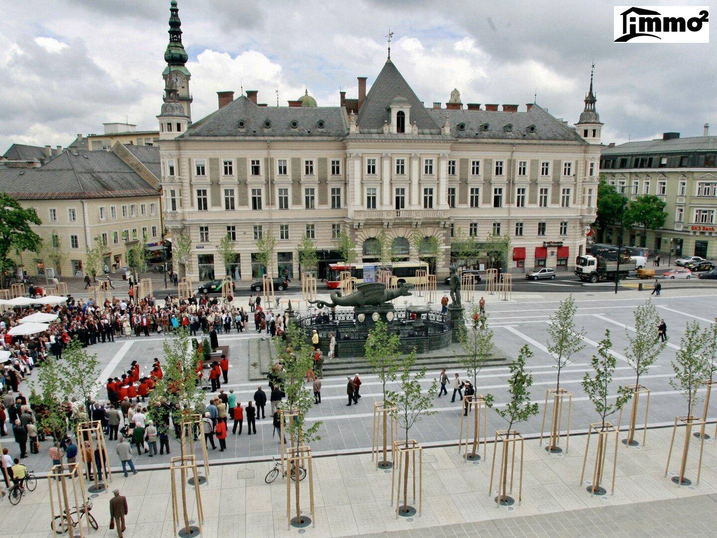 Historischer Stadtplatz mit einem großen Gebäude, Brunnen und vielen Menschen, umgeben von Bäumen.