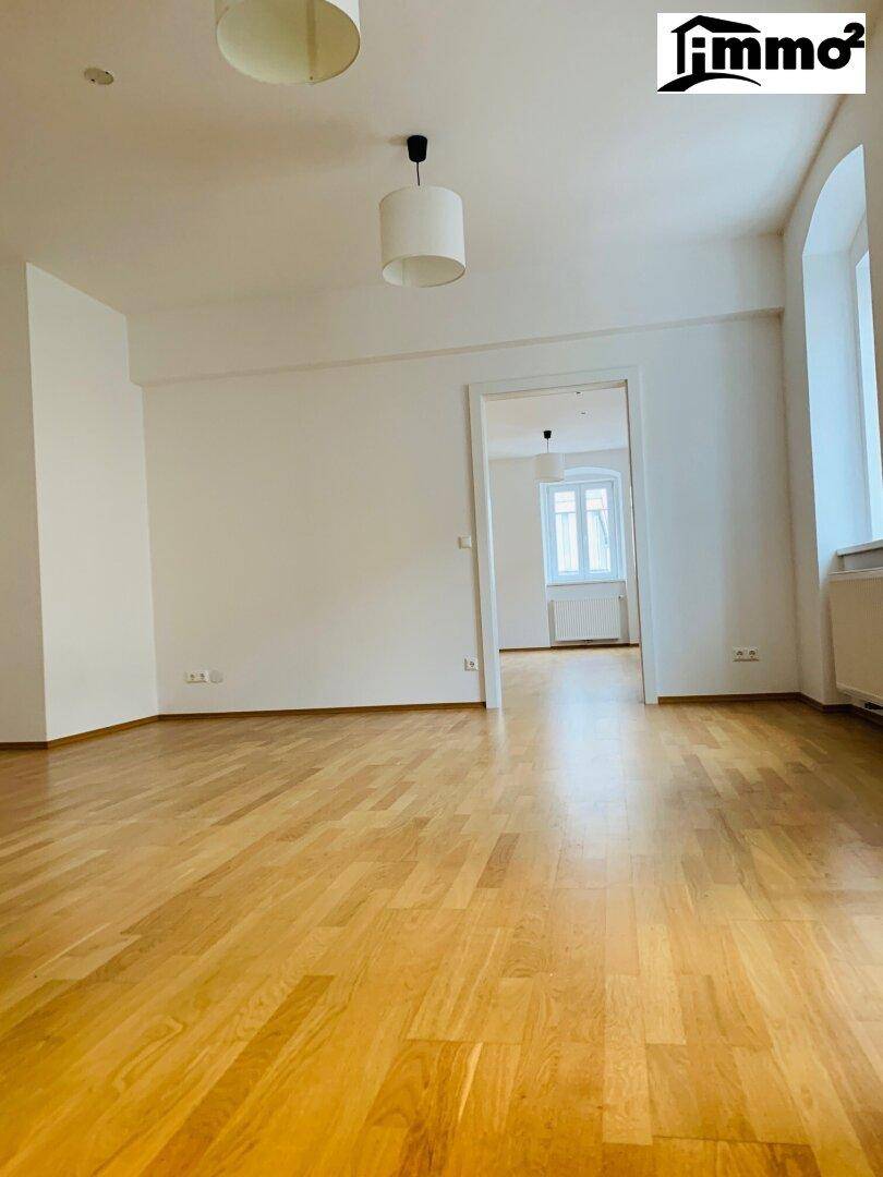 Heller, leerer Raum mit glänzendem Holzfußboden und weißen Wänden, mit Blick in einen weiteren Raum.