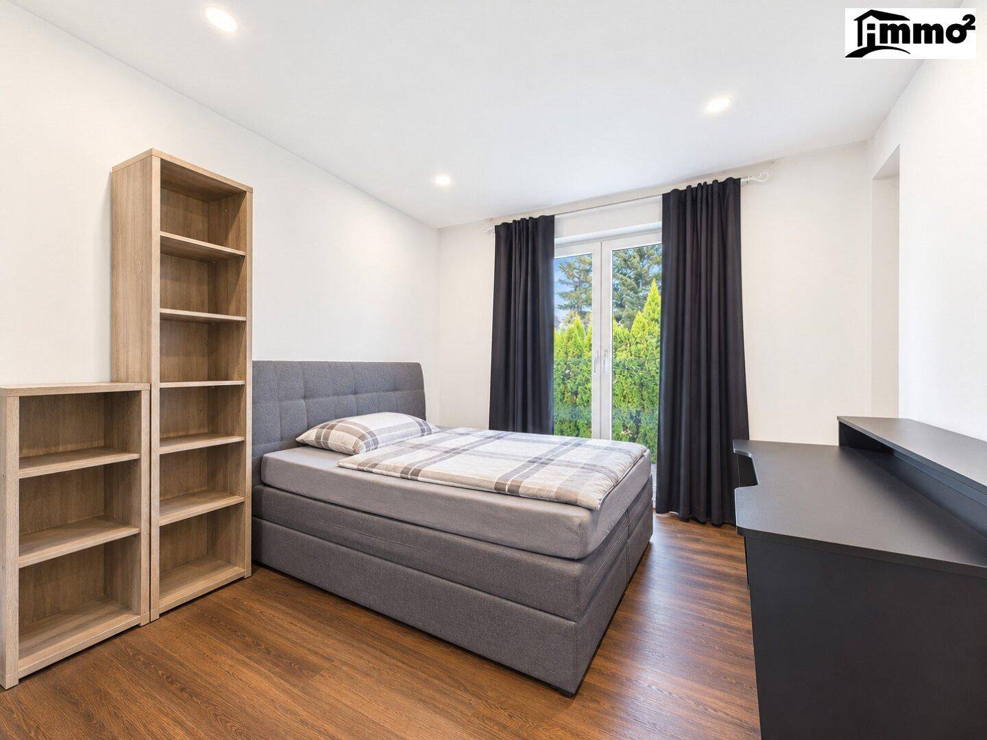 Schlafzimmer mit Einzelbett, Holzboden, Bücherregalen und Fenster mit dunklen Vorhängen.