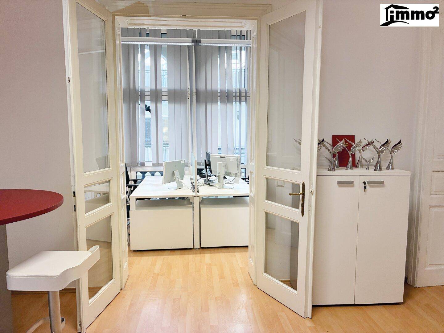 Blick durch Glastüren in ein helles Büro mit mehreren Arbeitsplätzen und Fensterfront.