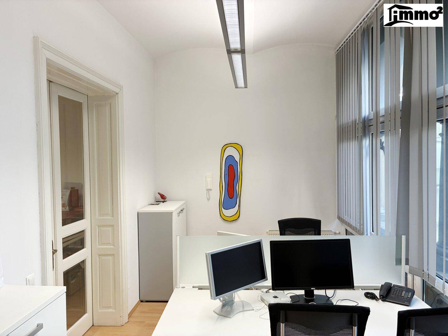 Modernes Büro mit mehreren Arbeitsplätzen, großen Fenstern und vertikalen Jalousien.