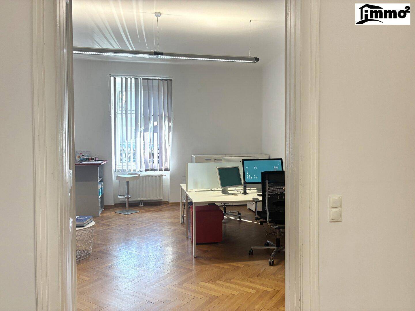 Großes Büro mit Fischgrätparkett, mehreren Arbeitsplätzen und Fenster mit Jalousien.
