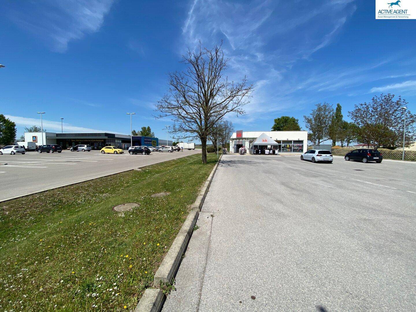 Weitläufiger Parkplatzbereich mit mehreren Geschäften im Hintergrund und grünen Bäumen.