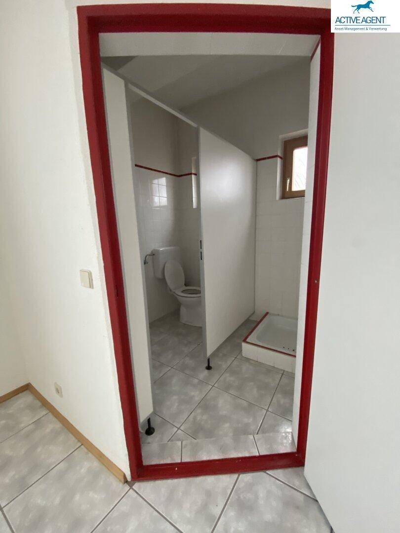 Kleiner Toilettenraum mit Fliesenboden, WC und Duschwanne, durch eine rote Türöffnung gesehen.