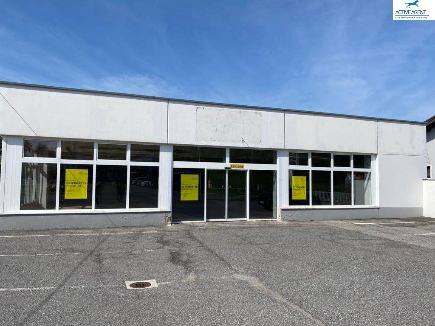 Frontansicht des Gewerbeobjekts mit breiten Schaufenstern und einem geräumigen Parkplatz, ideal für Einzelhandel.