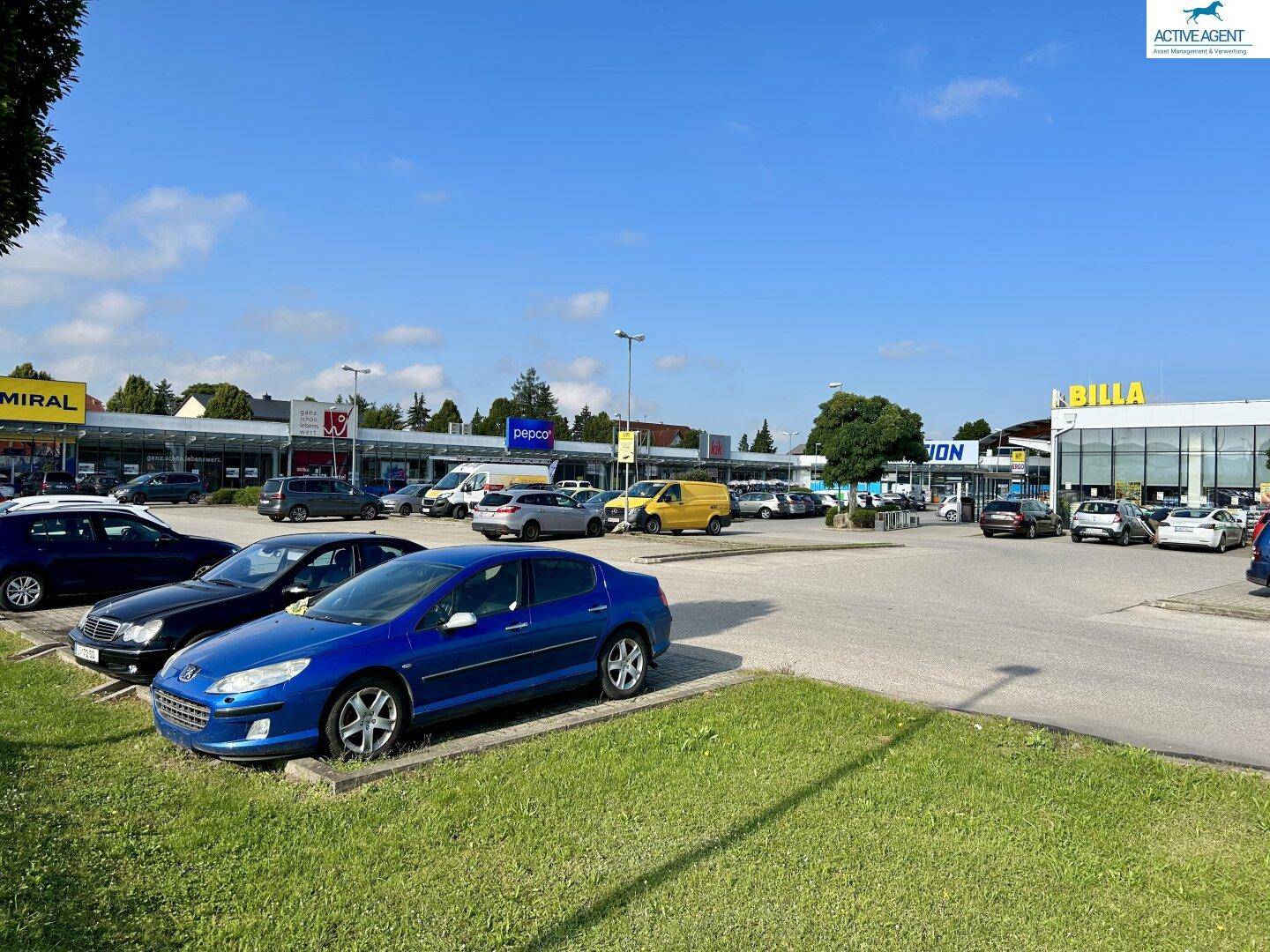 Weitläufiger Parkplatz eines Einkaufszentrums mit verschiedenen Geschäften und geparkten Fahrzeugen unter blauem Himmel.