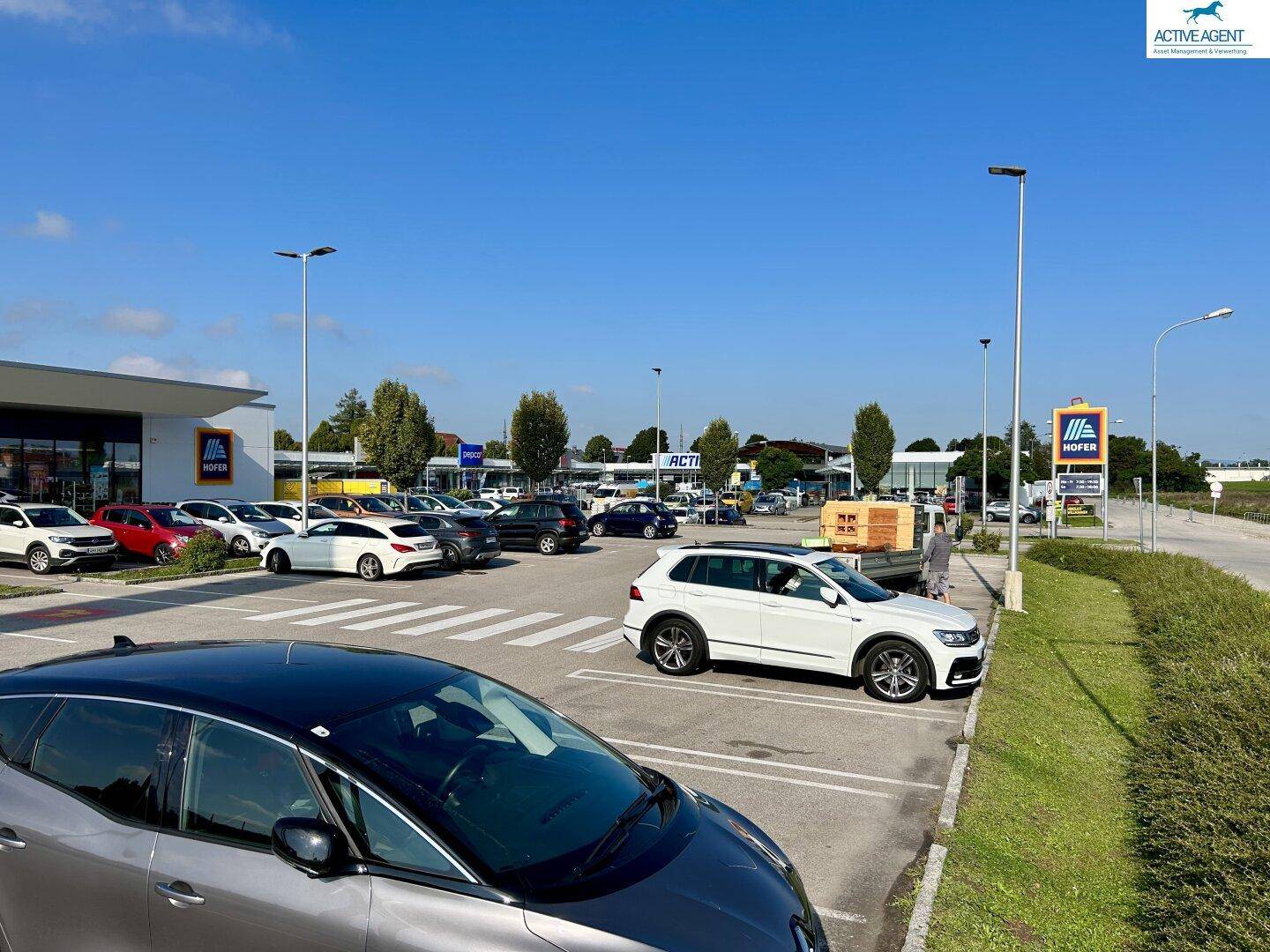 Großer Parkplatz vor einem Einkaufszentrum mit vielen geparkten Autos und einem Hofer-Schild.