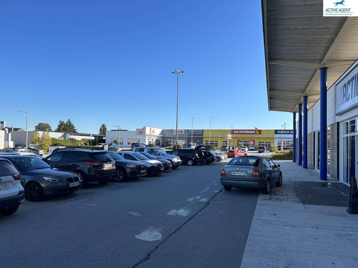 Ansicht des Fachmarktzentrums vom Parkplatz aus, mit einer Reihe von Geschäften und vielen geparkten Fahrzeugen.
