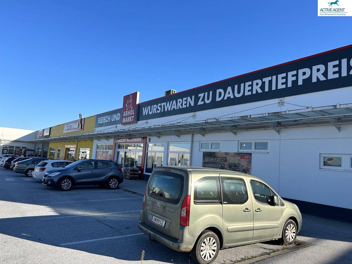 Außenansicht eines Einkaufszentrums mit verschiedenen Geschäften und einem belebten Parkplatz unter blauem Himmel.