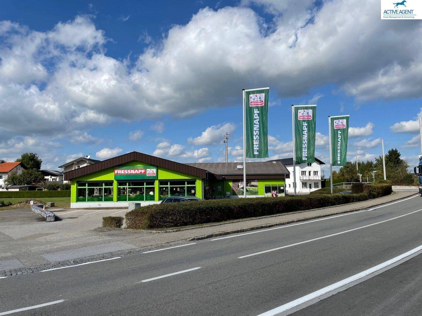 Ansicht der Gewerbeimmobilie von der Straße, mit grüner Fassade, Parkplätzen und Werbefahnen unter bewölktem Himmel.