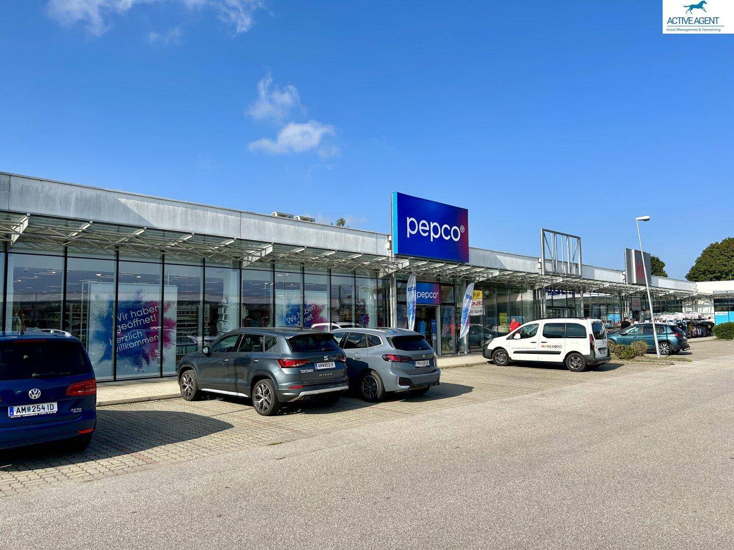 Moderne Ladenfront eines Geschäfts mit großen Fenstern und davor geparkten Autos auf einem weitläufigen Parkplatz.