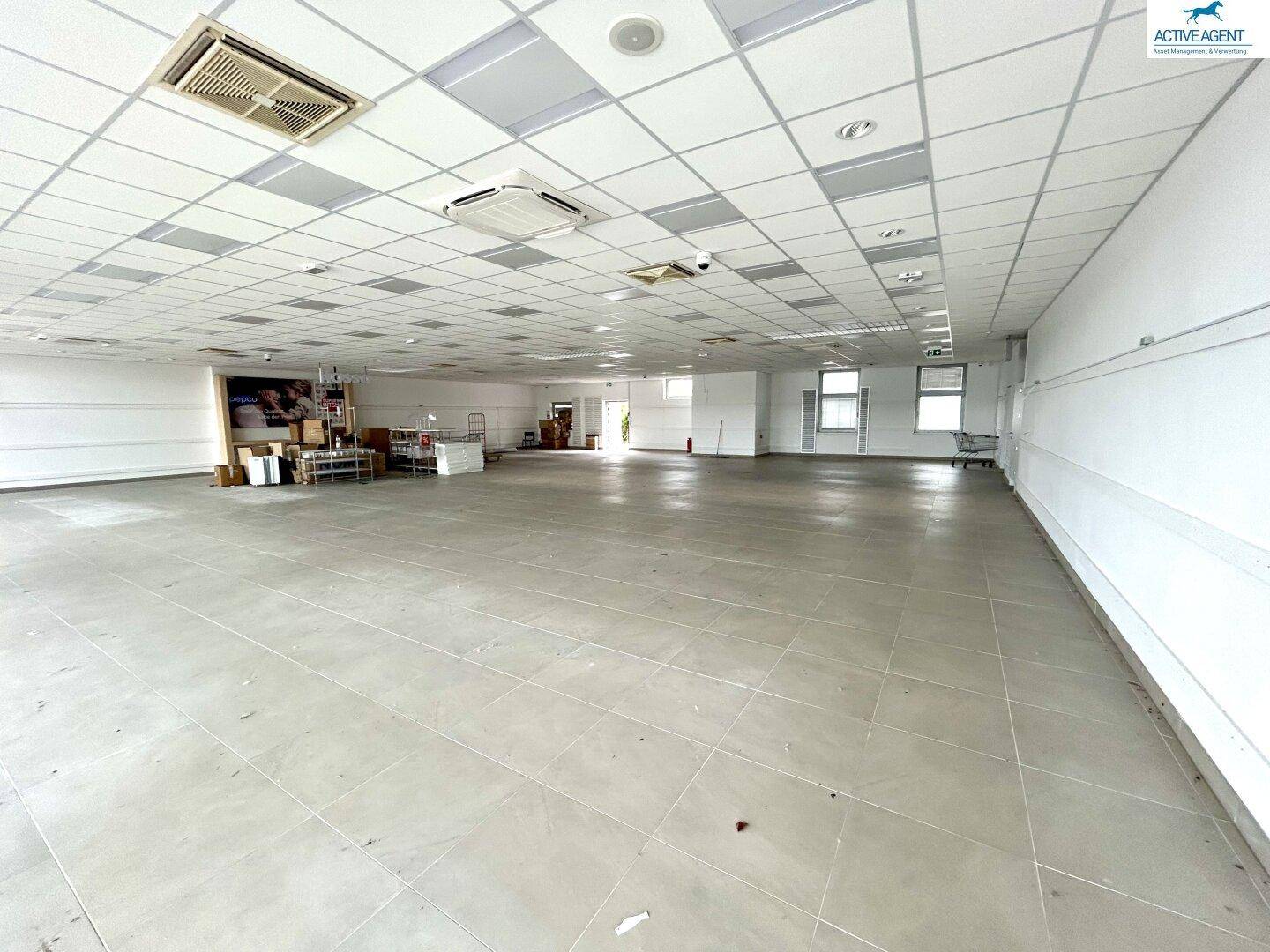 Weitläufiger, leerer Gewerberaum mit Fliesenboden, weißen Wänden und einer Decke mit integrierter Beleuchtung.