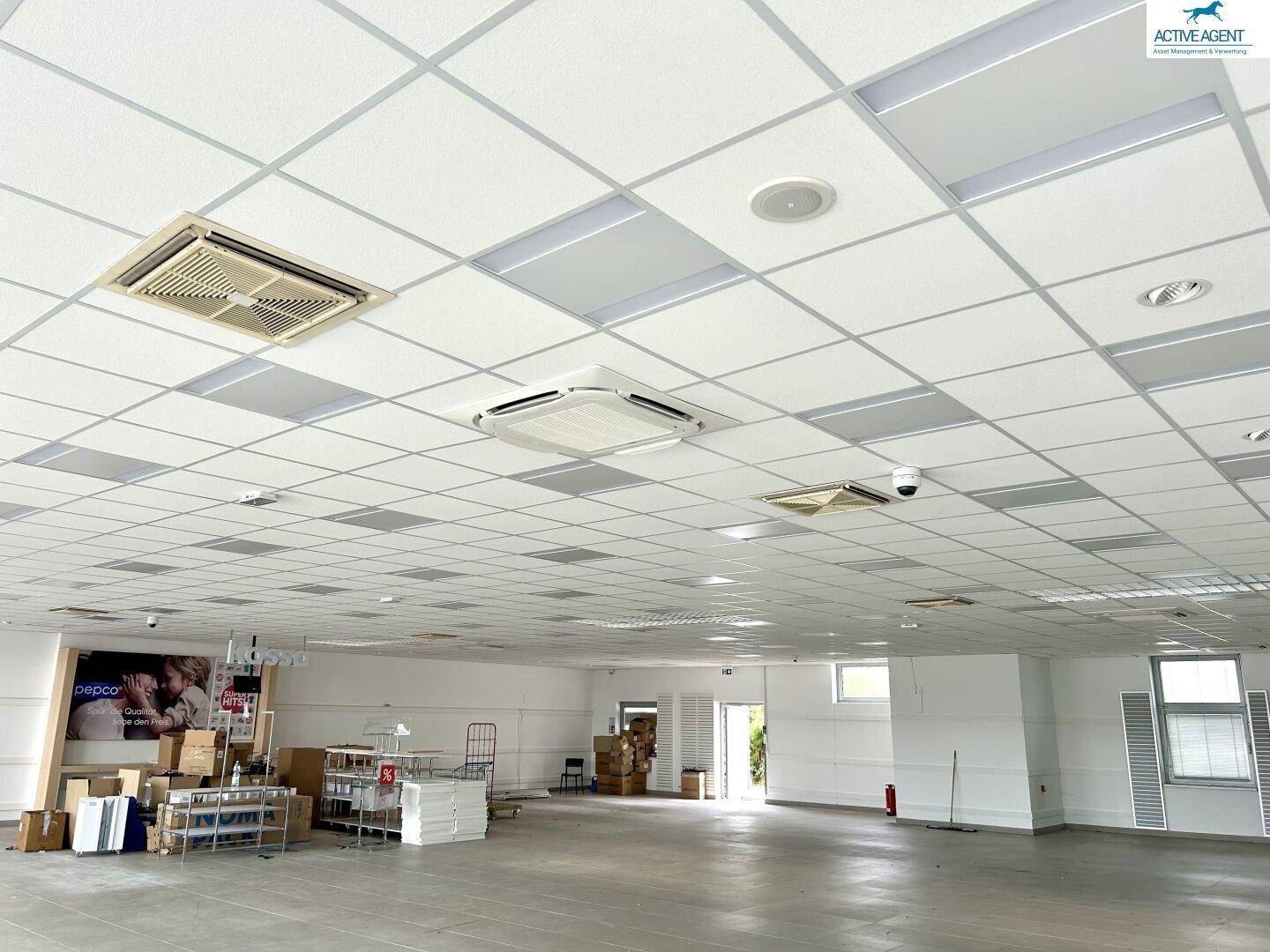 Geräumiger, leerer Ladenbereich mit Fliesenboden, Deckenplatten und sichtbaren Klimaanlagen und Kameras.