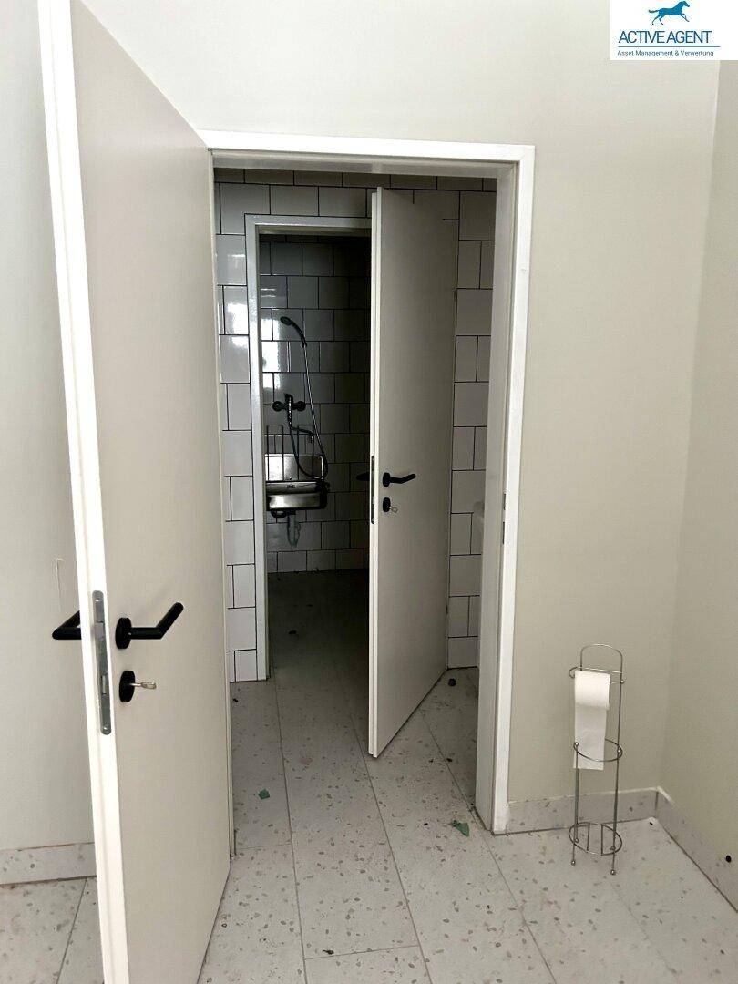 Einfacher Sanitärbereich mit gefliesten Wänden, einer Dusche und einer Toilettenpapierhalterung, die Renovierung benötigt.