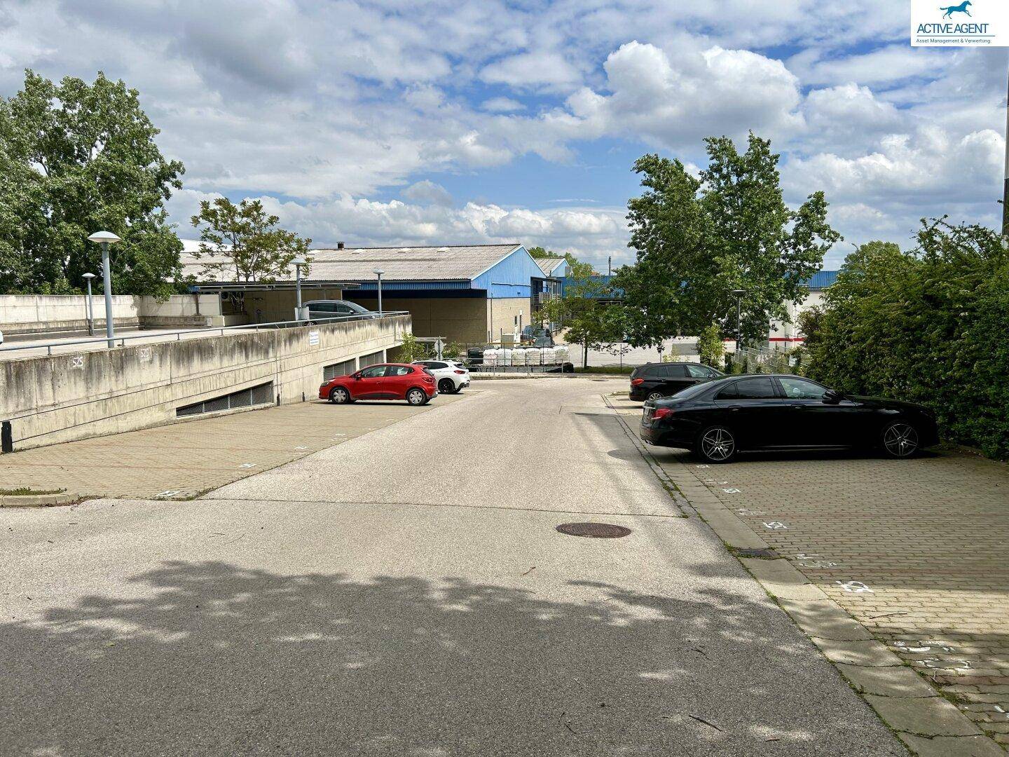 Außenparkplatz mit mehreren geparkten Fahrzeugen und einer Rampe zur Tiefgarage, umgeben von Grünflächen.