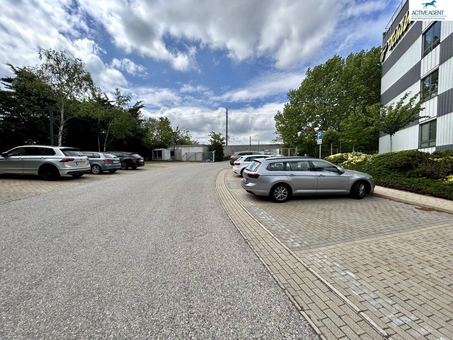 Außenparkplatz neben dem Bürogebäude mit mehreren geparkten Autos und Bäumen im Hintergrund unter blauem Himmel.