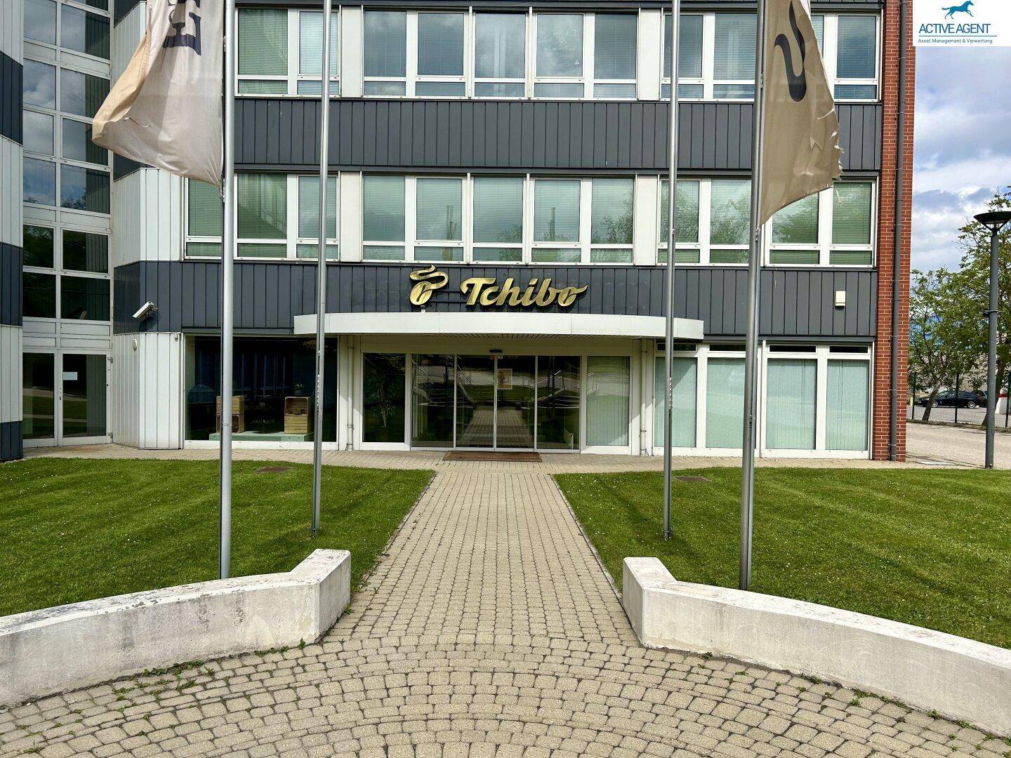 Eingangsbereich eines modernen Bürogebäudes mit gepflegtem Rasen und dem Tchibo-Logo über dem Eingang.