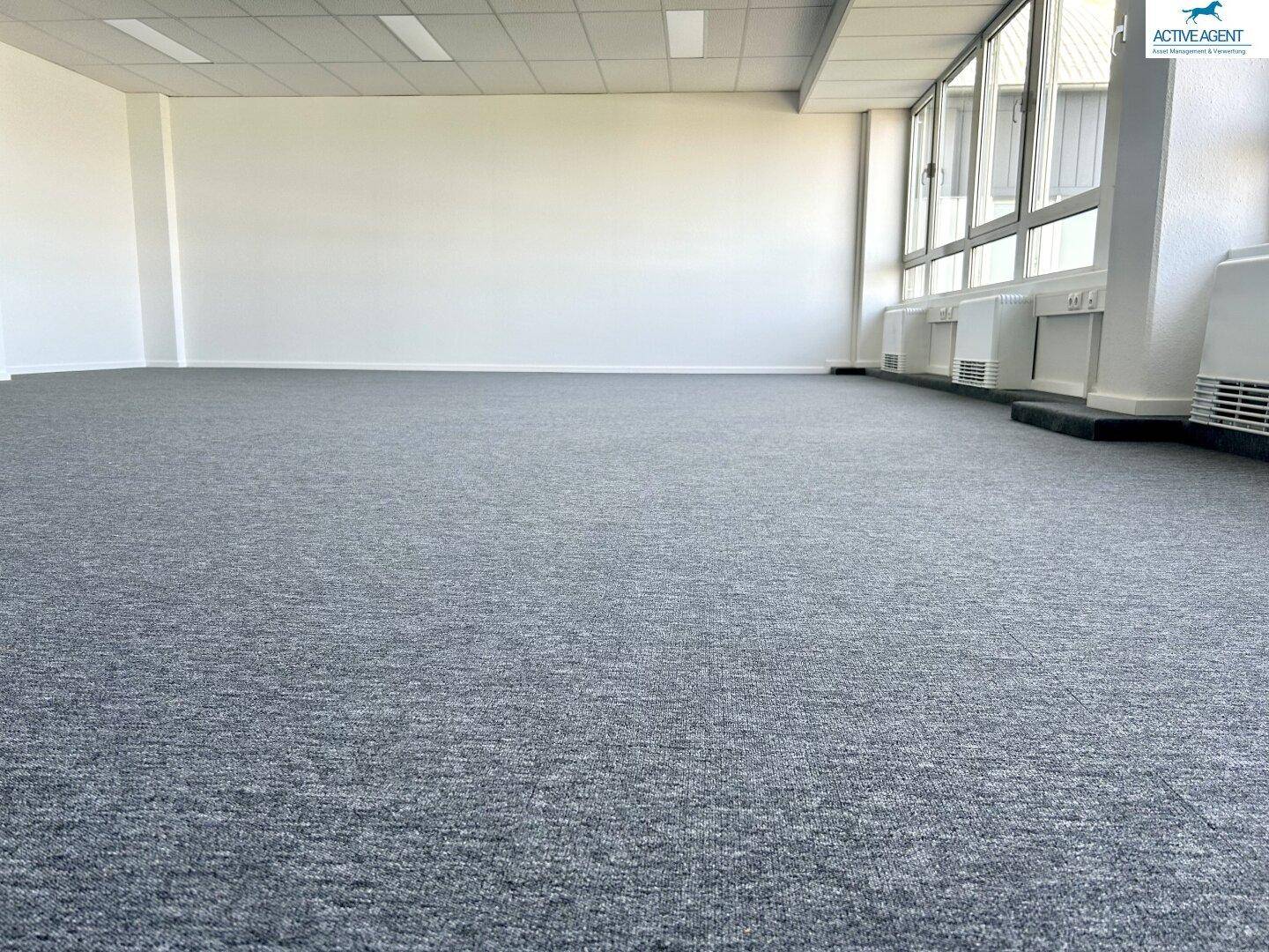 Weitläufiger Büroraum mit durchgehendem grauem Teppichboden und hellen Wänden, bereit zur Einrichtung.