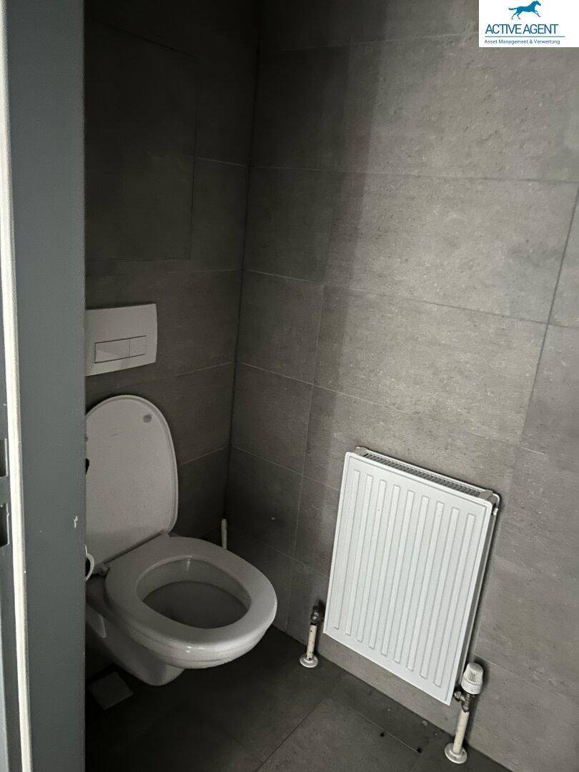 Separate Toilette mit Wand-WC, Heizkörper und durchgehend grauen Fliesen an Wänden und Boden.