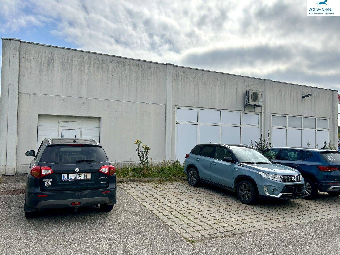 Rückseite eines Geschäftsgebäudes mit schlichter grauer Fassade, geparkten Autos und einer Klimaanlage an der Wand.