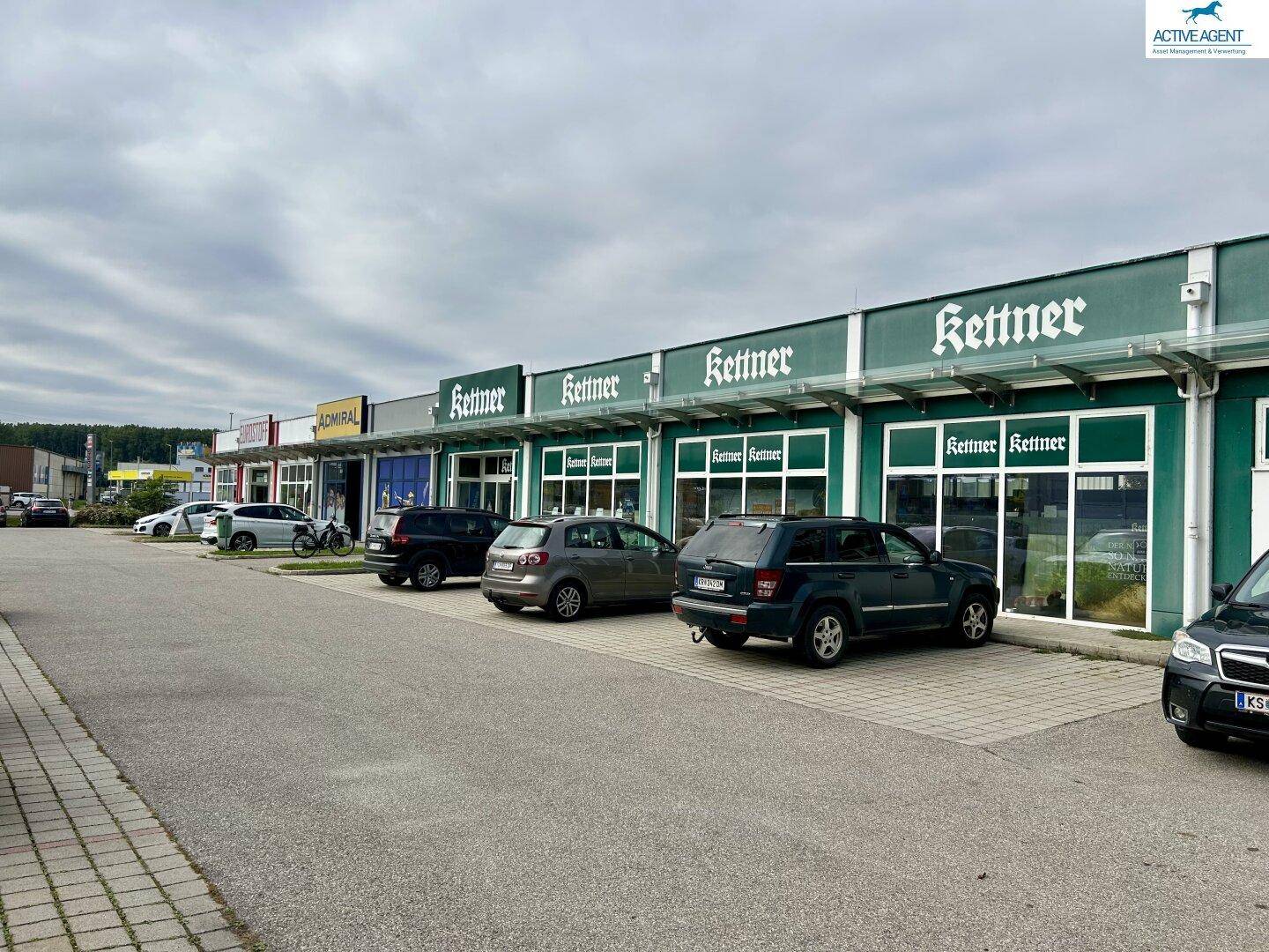 Reihe von Geschäftsgebäuden, darunter "Kettner" und "Admiral", mit einem großen Parkplatz davor unter bewölktem Himmel.