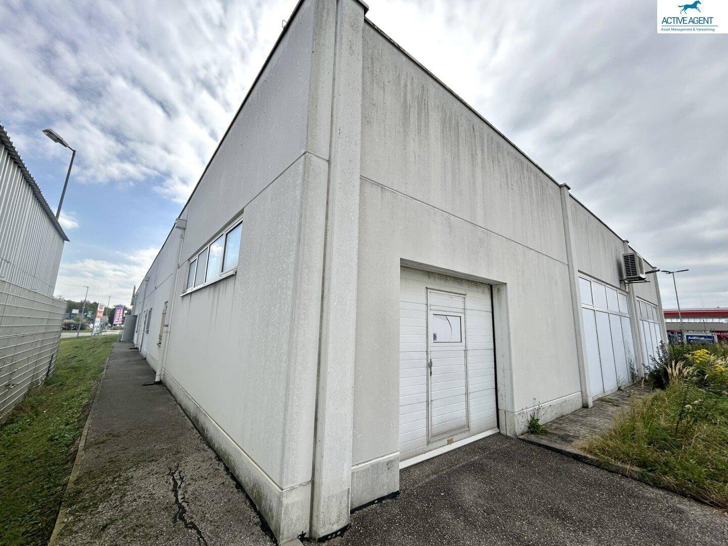 Seitenansicht eines Geschäftsgebäudes mit einfacher grauer Fassade, einer kleinen Tür und einigen Fenstern unter bewölktem Himmel.