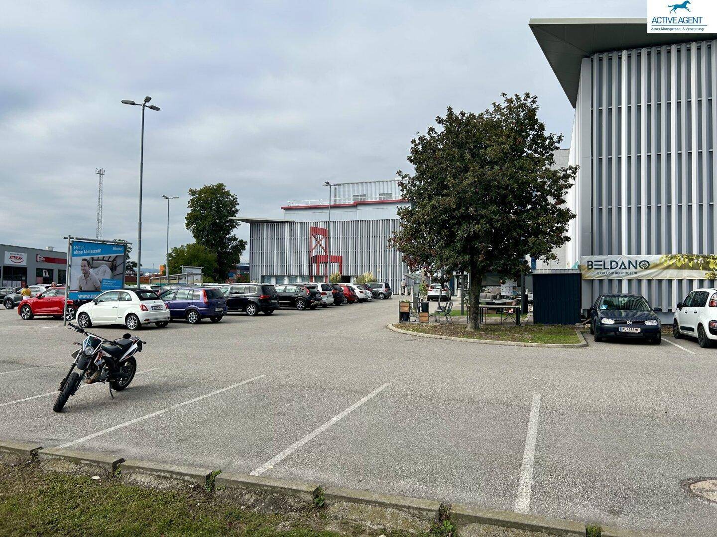 Großer Parkplatz mit verschiedenen Fahrzeugen und Motorrad, umgeben von Geschäftsgebäuden im Hintergrund unter bewölktem Himmel.