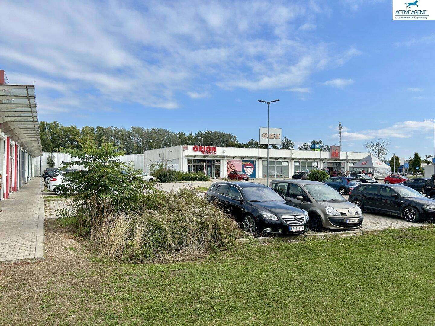 Weitere Außenansicht von Geschäftsgebäuden, darunter "ORION", mit Parkplatz und etwas verwachsenem Grün unter bewölktem Himmel.