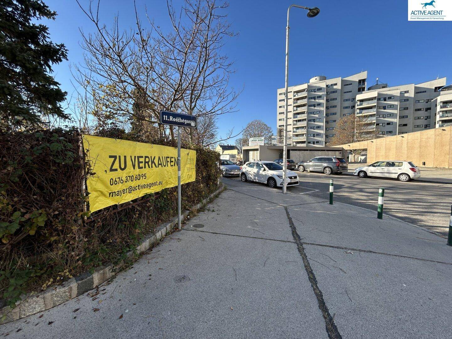 Straßenansicht mit geparkten Fahrzeugen, Wohngebäuden und einem 'Zu Verkaufen'-Banner an der Roschegasse.