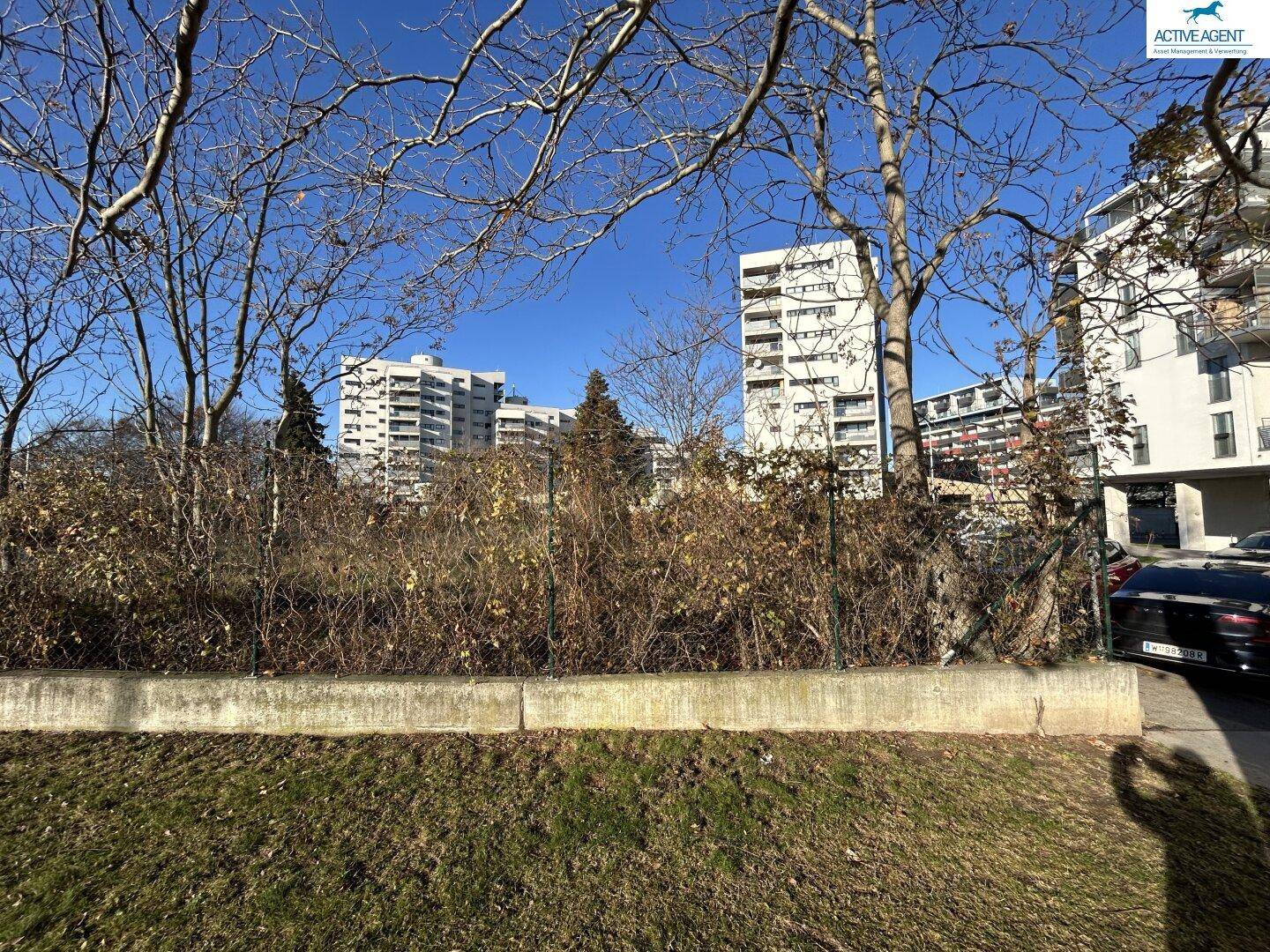 Blick über ein unbebautes Grundstück auf Wohngebäude in der Ferne unter blauem Himmel.