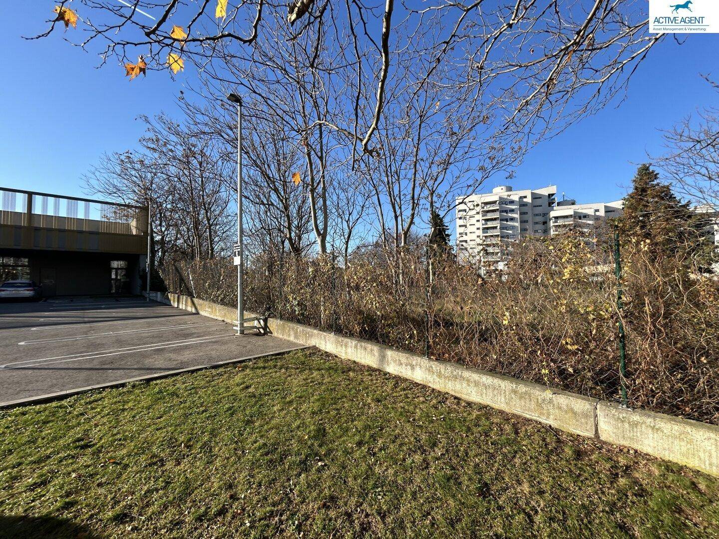 Unbebautes Grundstück neben einem Parkplatz und einer Brücke mit Gebäuden im Hintergrund.