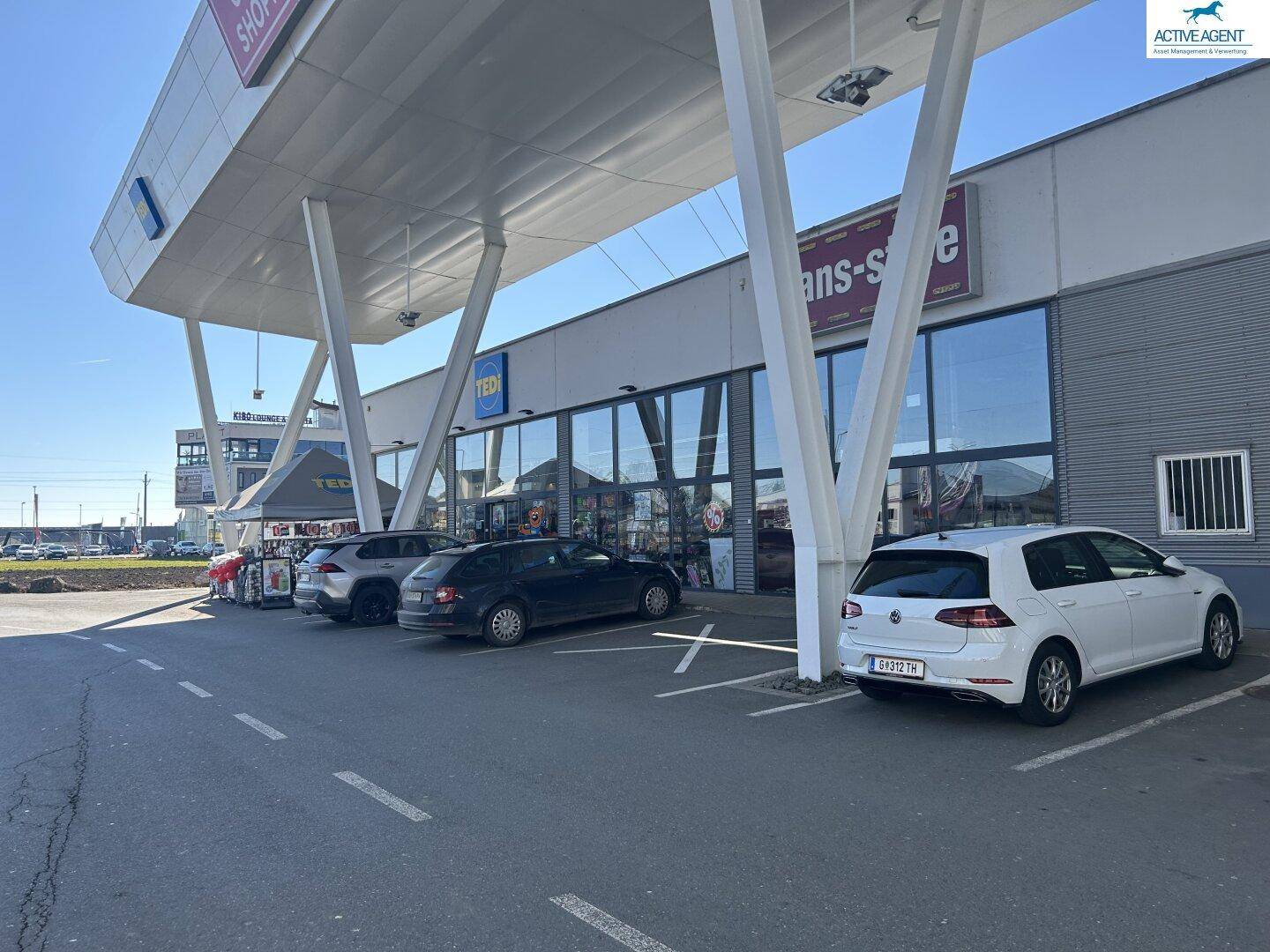 Seitenansicht eines Einkaufszentrums mit Geschäften, großem Vordach und geparkten Autos auf dem Parkplatz.