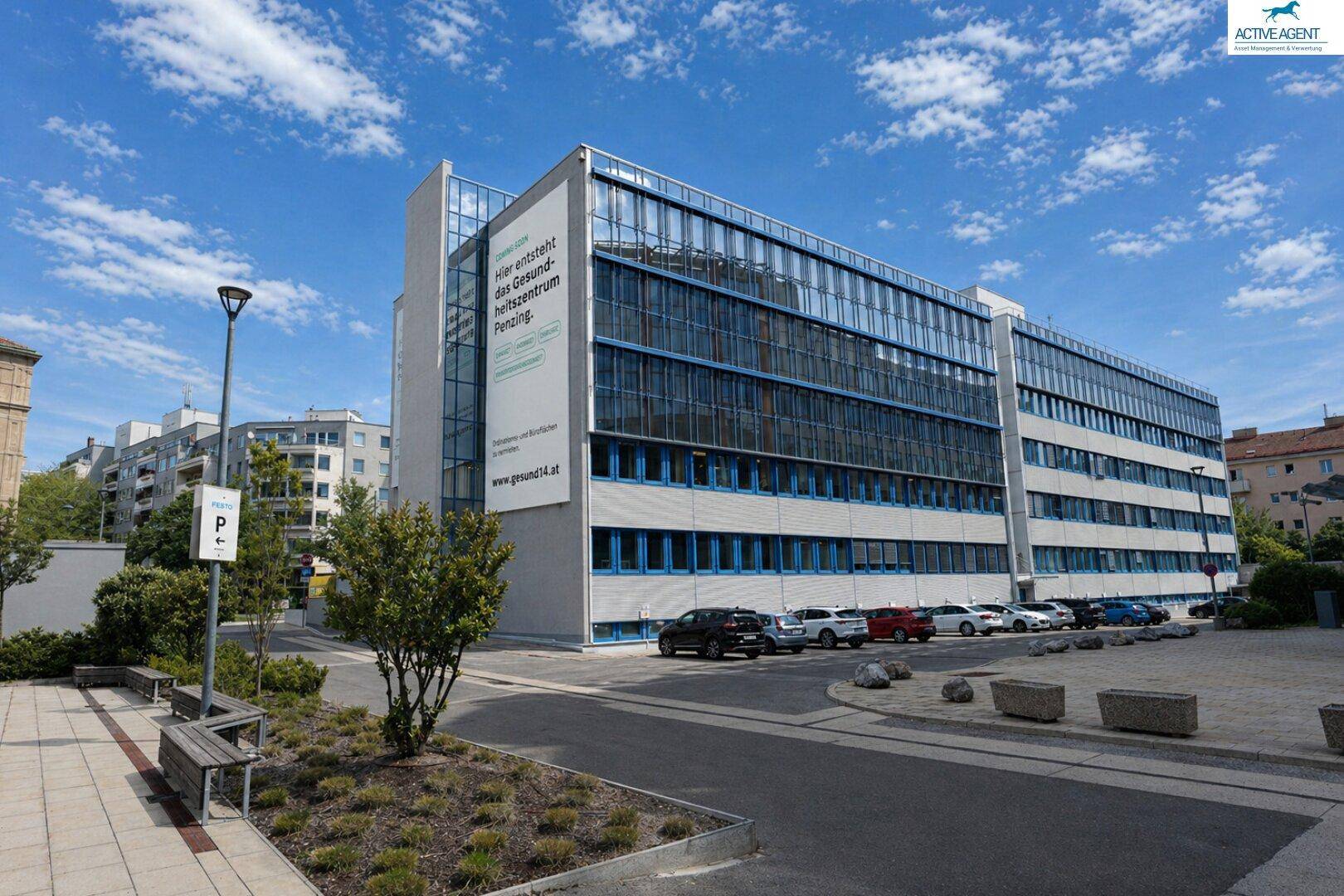 Modernes Geschäftsgebäude mit Glasfronten und Hinweisschildern für ein zukünftiges Gesundheitszentrum, umgeben von Grünflächen.