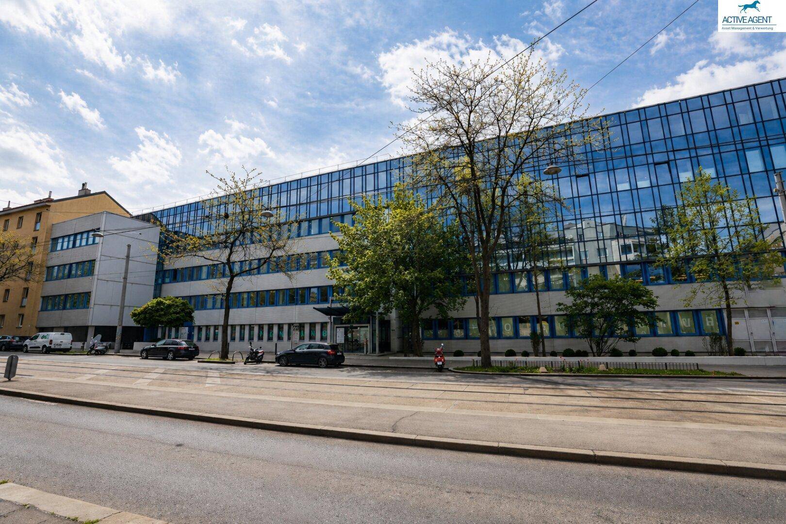 Lange Ansicht eines modernen Bürogebäudes mit durchgehender Glasfassade entlang einer belebten Straße mit Bäumen.
