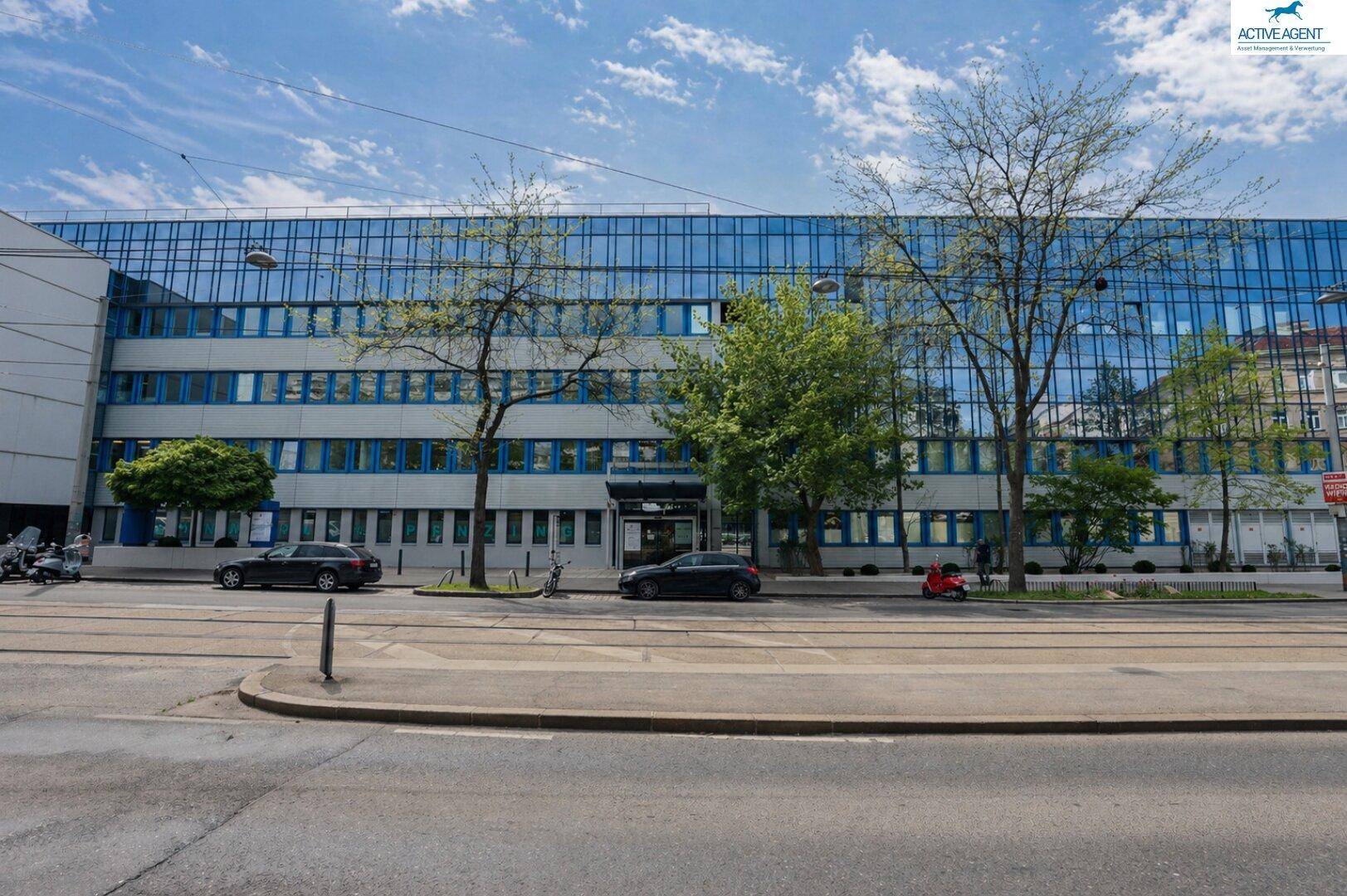 Weitläufige Straßenansicht eines modernen Bürogebäudes mit durchgehender Glasfassade und Bäumen im Vordergrund.