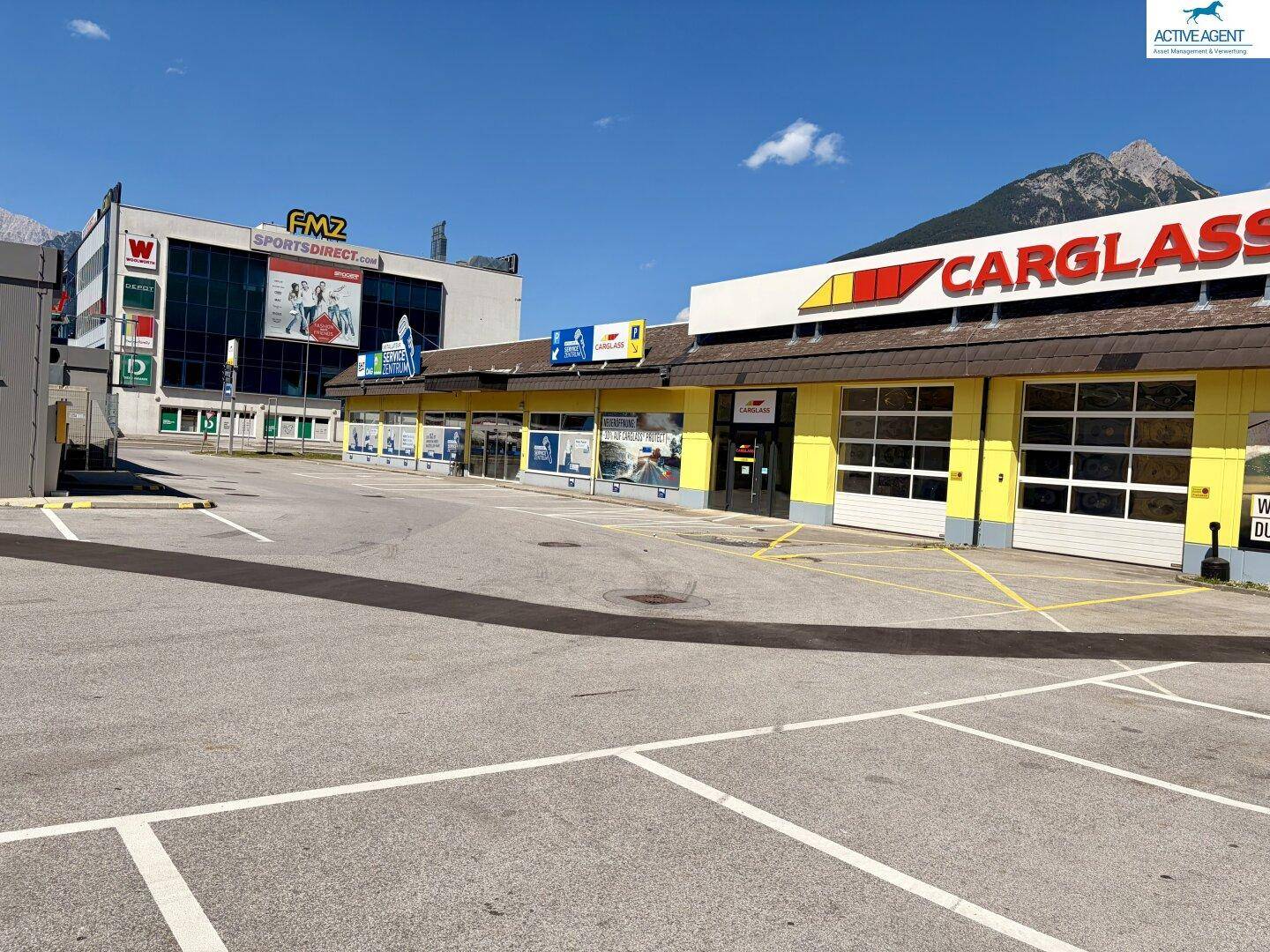 Weitläufiger Parkplatz vor dem gelben Geschäftsgebäude und einem größeren Einkaufszentrum im Hintergrund.