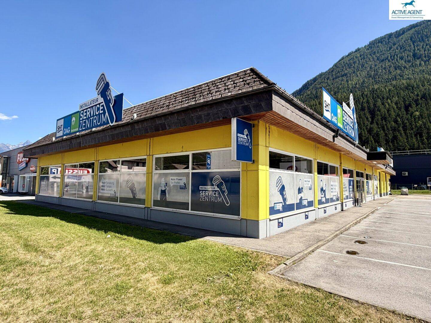 Seitenansicht des gelben Geschäftsgebäudes mit Parkplätzen und grüner Wiese im Vordergrund, umgeben von Bergen.