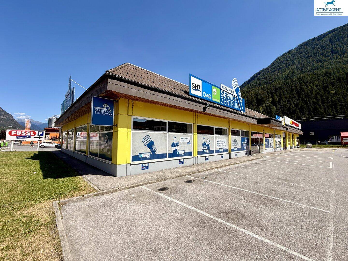 Ansicht des gelben Geschäftsgebäudes mit Werbeschildern und einem großen Parkplatz unter blauem Himmel.