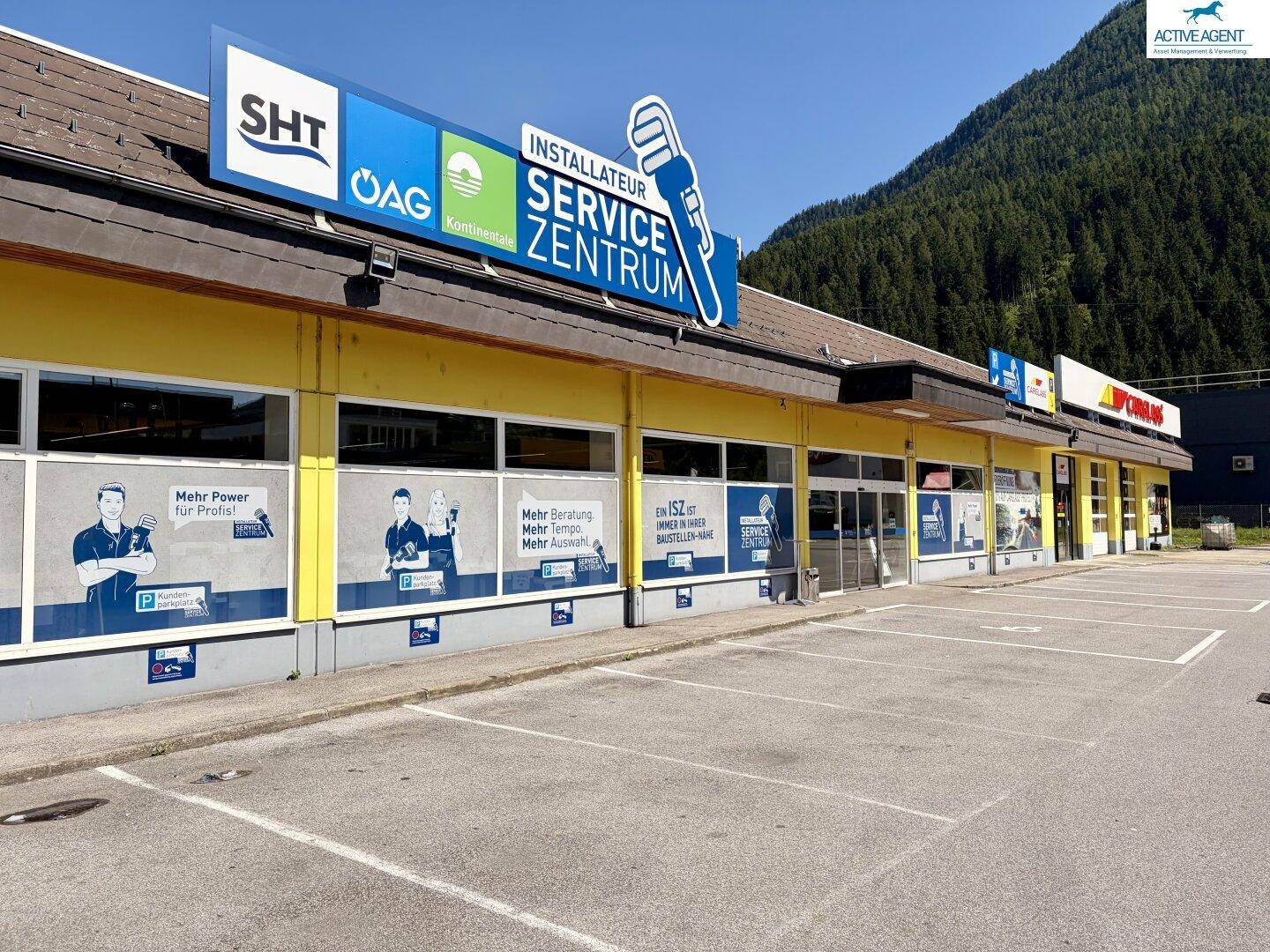 Frontansicht des gelben Servicezentrums mit Werbeschildern und leeren Parkplätzen an einem sonnigen Tag.