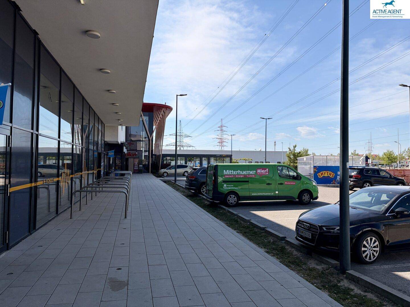 Seitenansicht eines modernen Geschäftsgebäudes mit Parkplätzen und Stromleitungen im Hintergrund unter blauem Himmel.