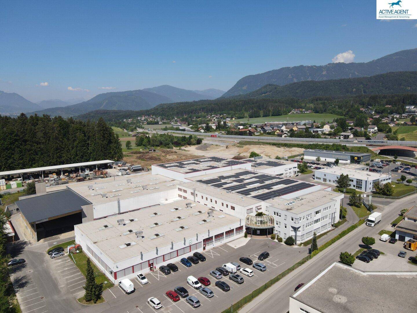 Luftaufnahme eines Gewerbekomplexes mit Solaranlagen auf den Dächern und beeindruckendem Bergblick im Hintergrund.