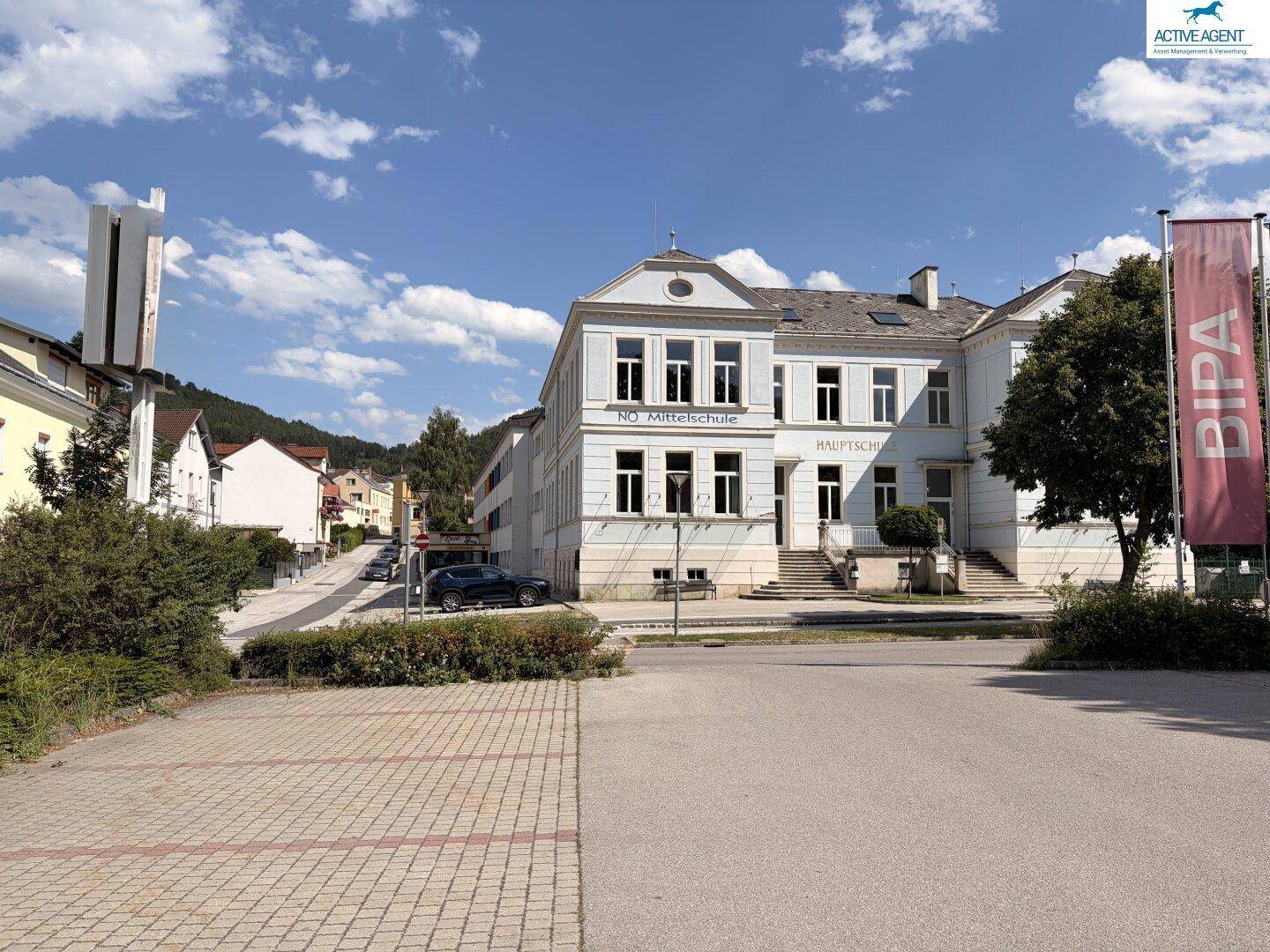 Historisches Schulgebäude mit klassischer Architektur, umgeben von einer städtischen Landschaft und Bergen im Hintergrund.