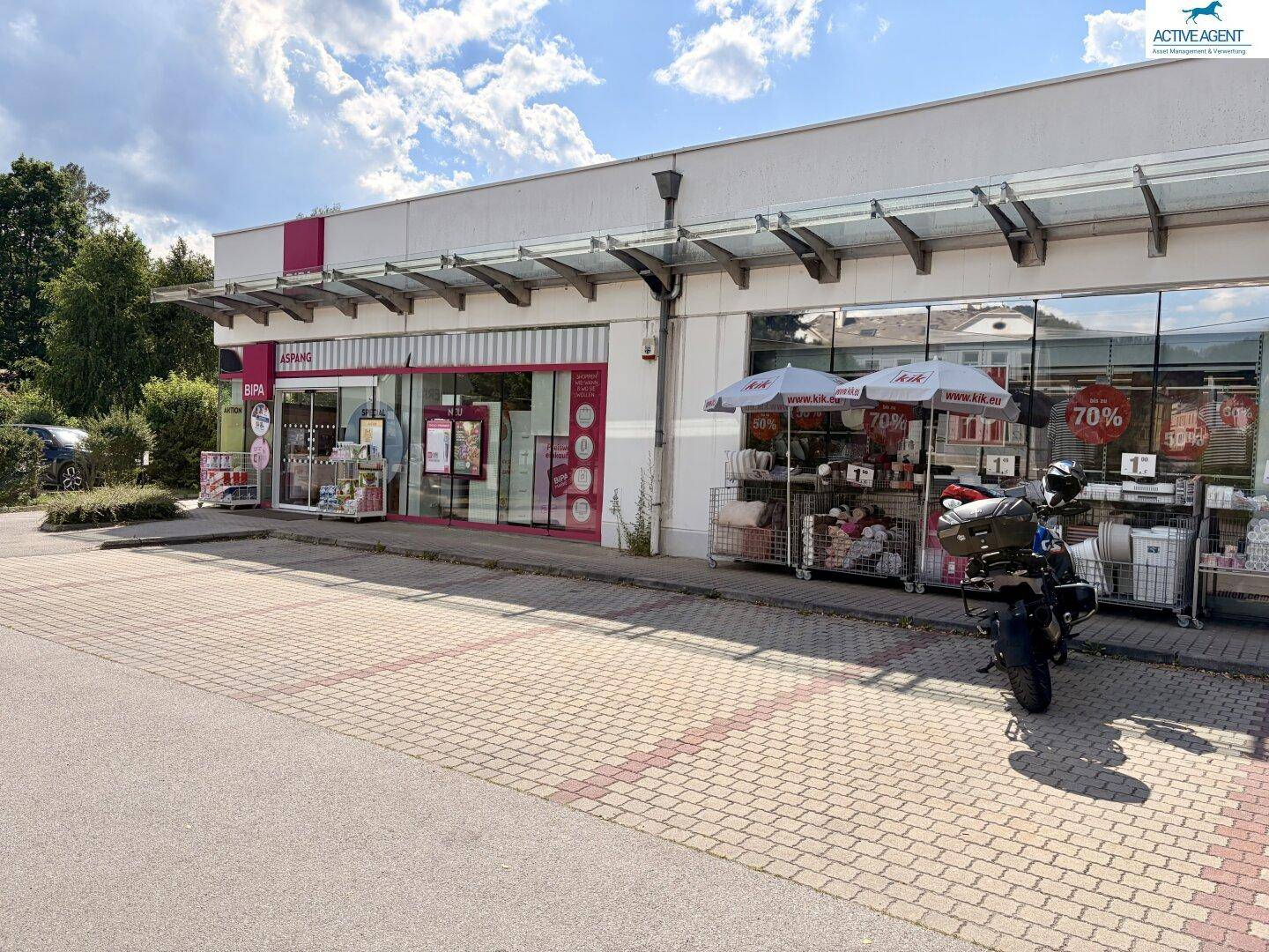Nahaufnahme der Geschäftsfassade mit Schaufenstern, Vordach und einem Motorrad auf dem gepflasterten Parkplatz.