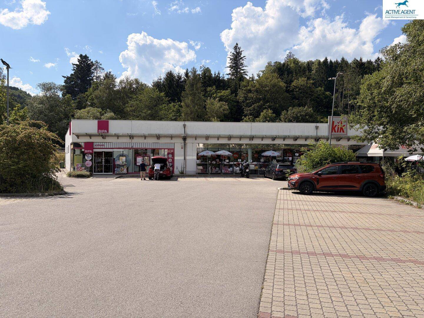 Gesamtansicht des Geschäftsgebäudes mit großem Parkplatz und bewaldeter Umgebung unter blauem Himmel.