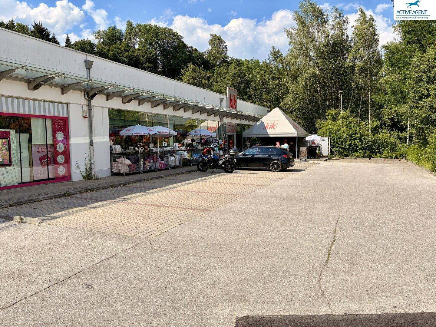 Seitenansicht des Geschäftsgebäudes mit Parkplatz, umgeben von Bäumen und viel Grün unter blauem Himmel.