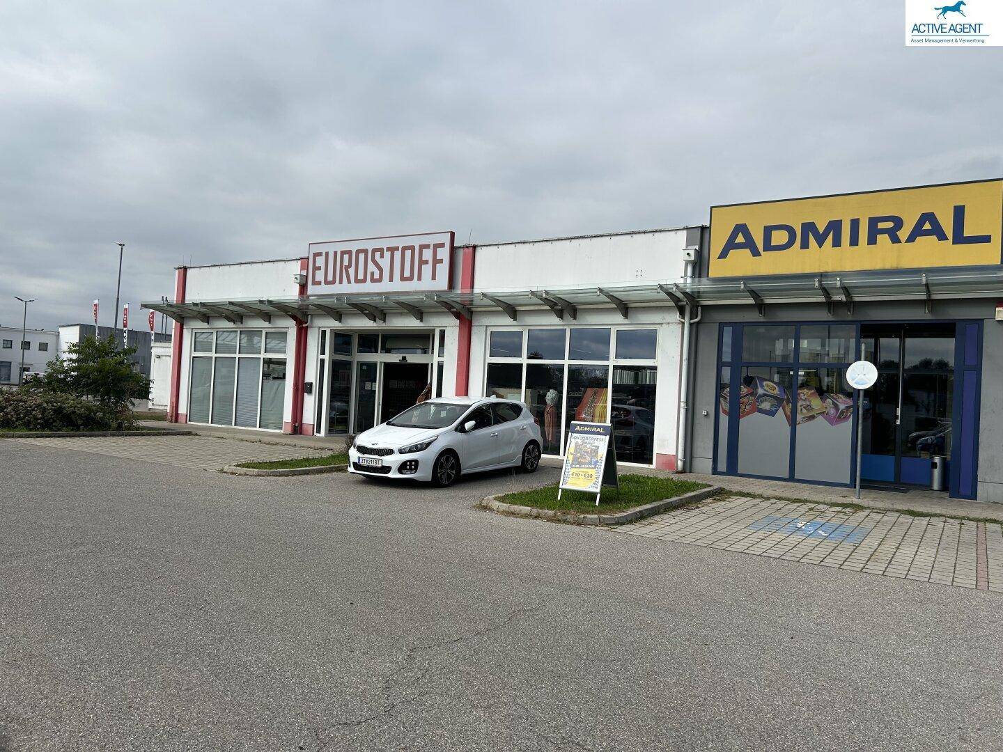 Außenansicht eines Gewerbegebäudes mit mehreren Ladenfronten und einem Parkplatz davor.