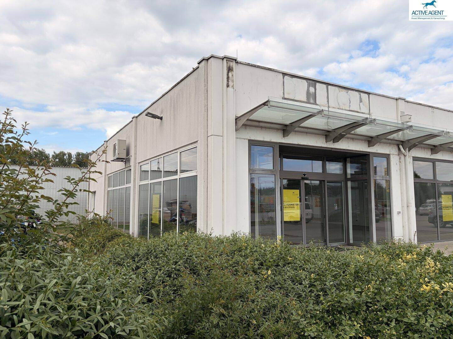 Eckansicht eines Gewerbegebäudes mit großen Fenstern, Büschen im Vordergrund und Klimaanlagen an der Fassade.