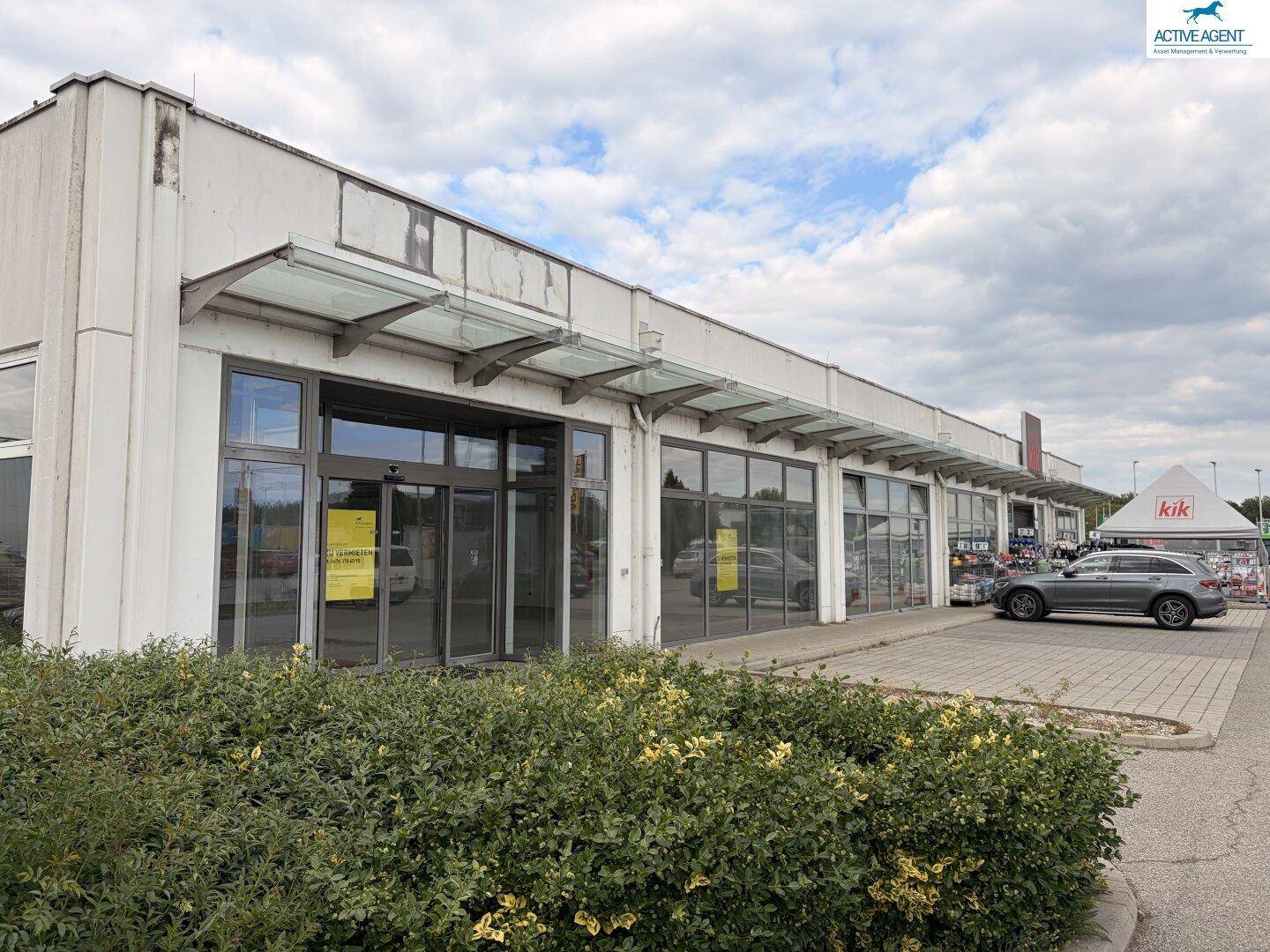 Lange Fassade eines Gewerbegebäudes mit mehreren Ladenfronten, Glasvordächern und Büschen im Vordergrund.