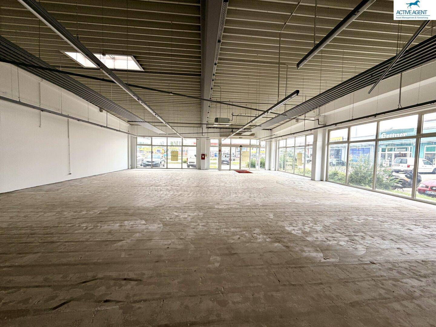 Großer, leerer Gewerberaum mit Betonboden, hohen Decken und großen Fenstern zur Außenseite.
