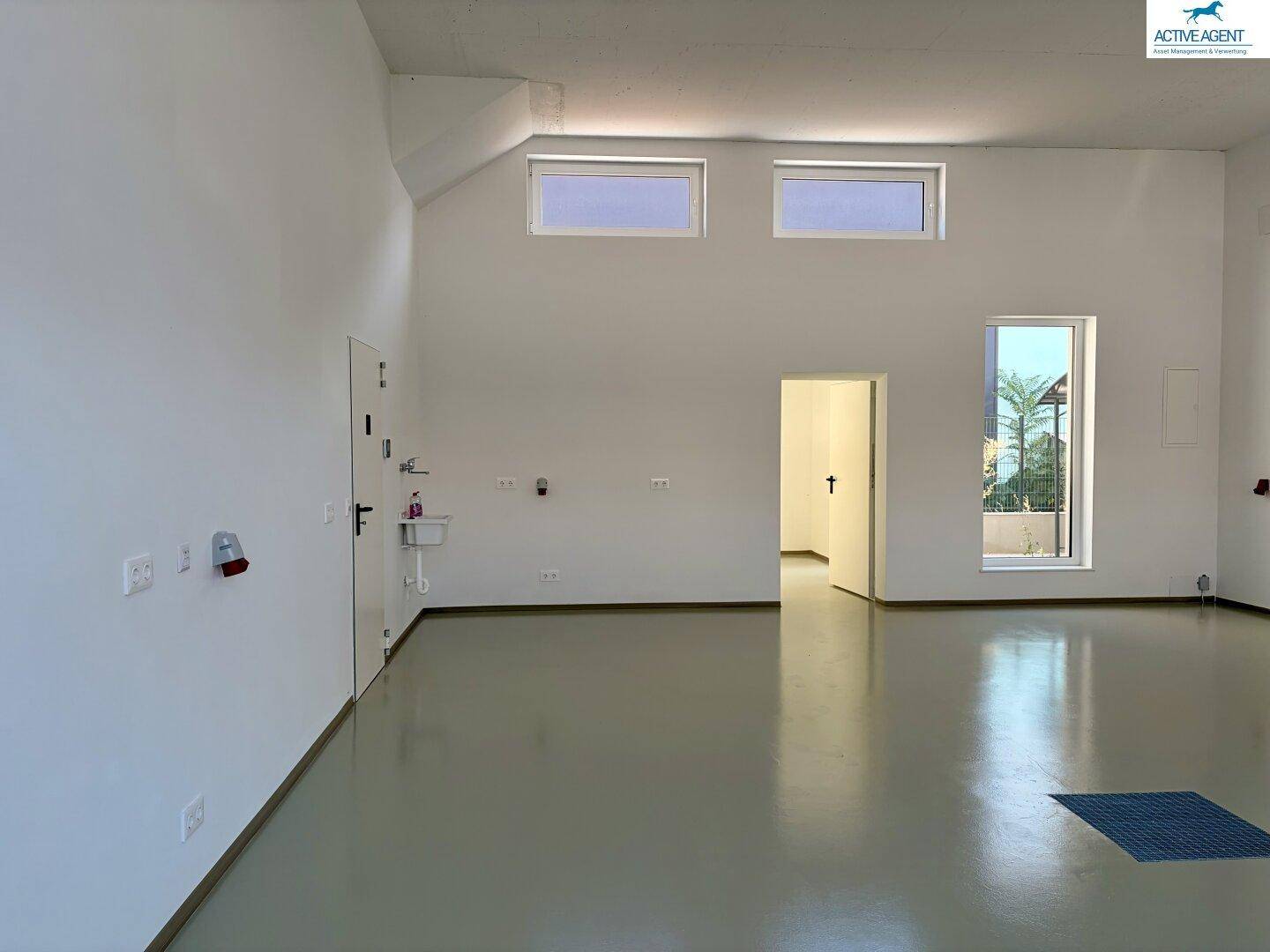 Heller, leerer Lagerraum mit hohen Decken, großen Fenstern und einem kleinen Waschbecken.